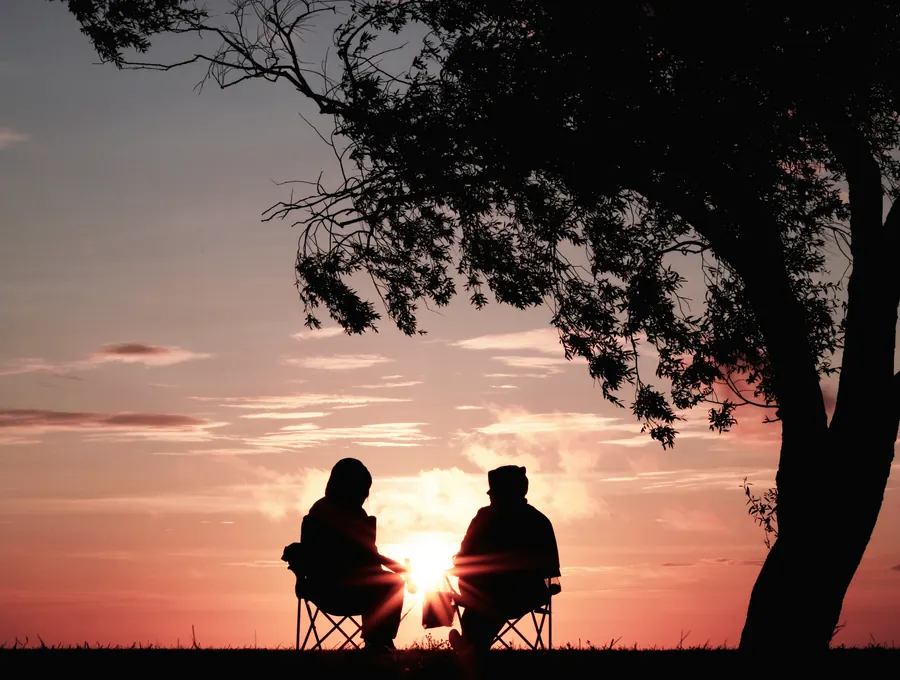 Two people sitting side by side in silhouette, talking as the sun sets