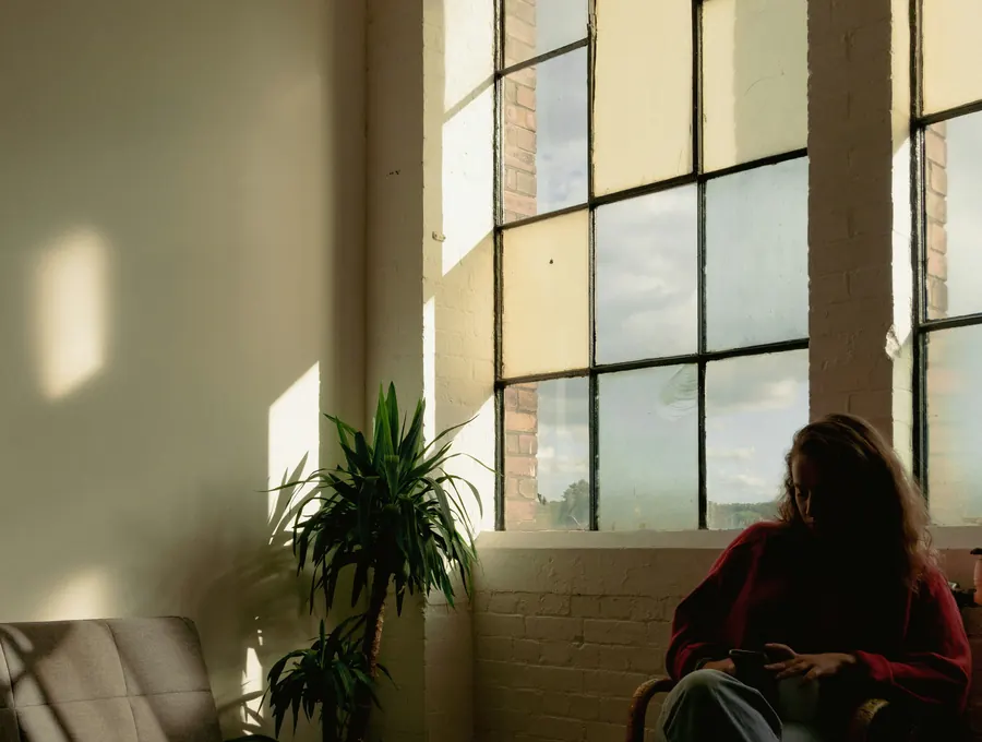 A person sitting quietly by a large window in soft, sheltered light, beside a plant